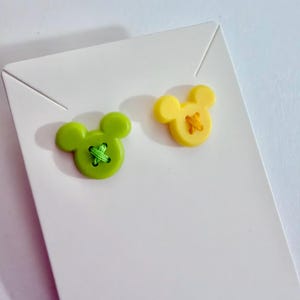 May include: Two Mickey Mouse-shaped buttons. One is lime green with green thread, the other is yellow with yellow thread. The buttons are on a white card, perfect for crafting or sewing projects. The buttons are made of plastic.