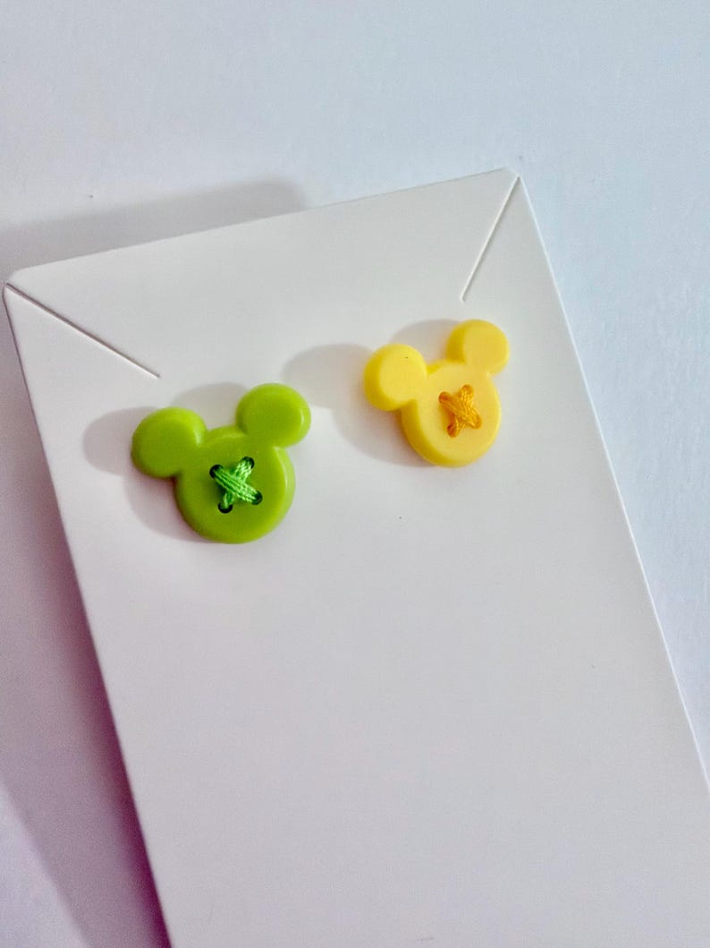 May include: Two Mickey Mouse-shaped buttons. One is lime green with green thread, the other is yellow with yellow thread. The buttons are on a white card, perfect for crafting or sewing projects. The buttons are made of plastic.