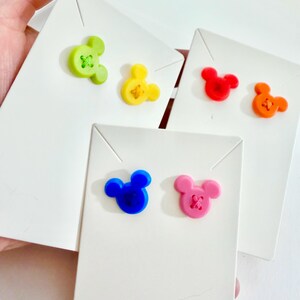 May include: A collection of colorful Mickey Mouse-shaped buttons in green, yellow, red, orange, blue, and pink, each attached to a white card. The buttons have a stitched design in the center.