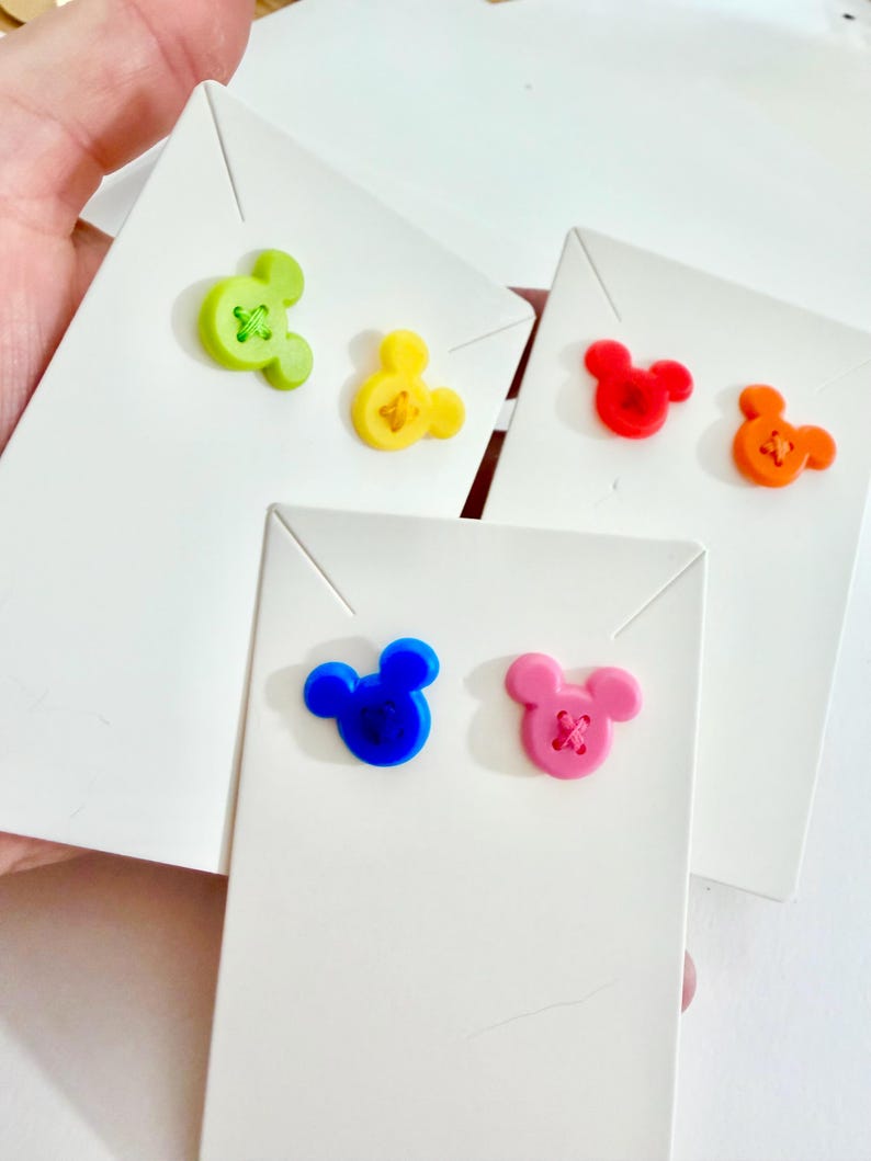 May include: A collection of colorful Mickey Mouse-shaped buttons in green, yellow, red, orange, blue, and pink, each attached to a white card. The buttons have a stitched design in the center.