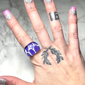 May include: A chunky, square-shaped ring with a vibrant purple base and a white giraffe-like pattern. The ring is made of a glossy material and is worn on a finger. The ring's design is eye-catching and modern.