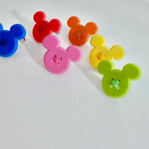 May include: Six colourful buttons in the shape of a mouse's head. The buttons are blue, red, orange, pink, yellow, and green, each with a contrasting thread detail. Suitable for sewing and craft projects.