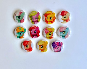 Può includere: Un set di undici spille rotonde con illustrazioni colorate di pony dei cartoni animati con vari colori di criniera, tra cui rosa, giallo, arancione, verde e viola, disposte in tre file su uno sfondo bianco uniforme.