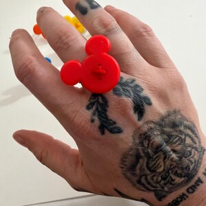 May include: A vibrant red Mickey Mouse-shaped ring made of plastic. The ring has a button-like detail in the centre. Other colourful rings are visible in the background. A playful accessory.