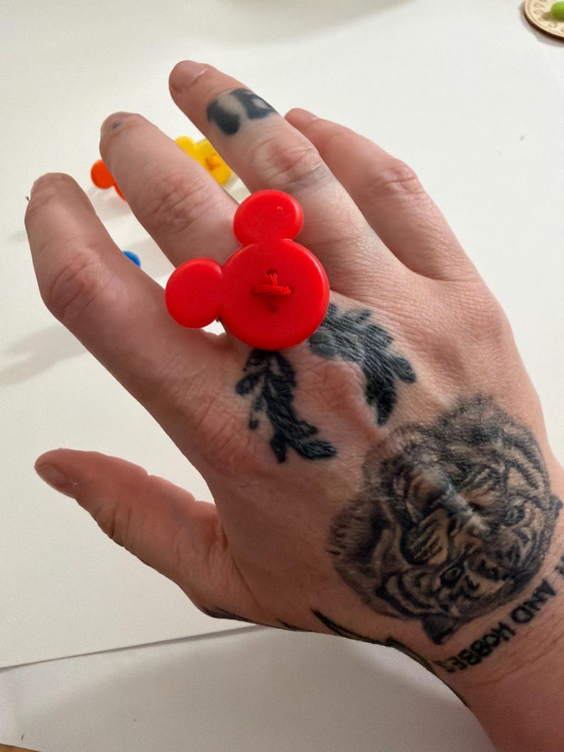 May include: A vibrant red Mickey Mouse-shaped ring made of plastic. The ring has a button-like detail in the centre. Other colourful rings are visible in the background. A playful accessory.