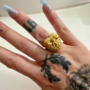 May include: A gold-colored heart-shaped ring with an embossed cherub design. The ring is worn on a finger. The ring's design features a heart shape with a cherub on top. The ring is a piece of jewelry.