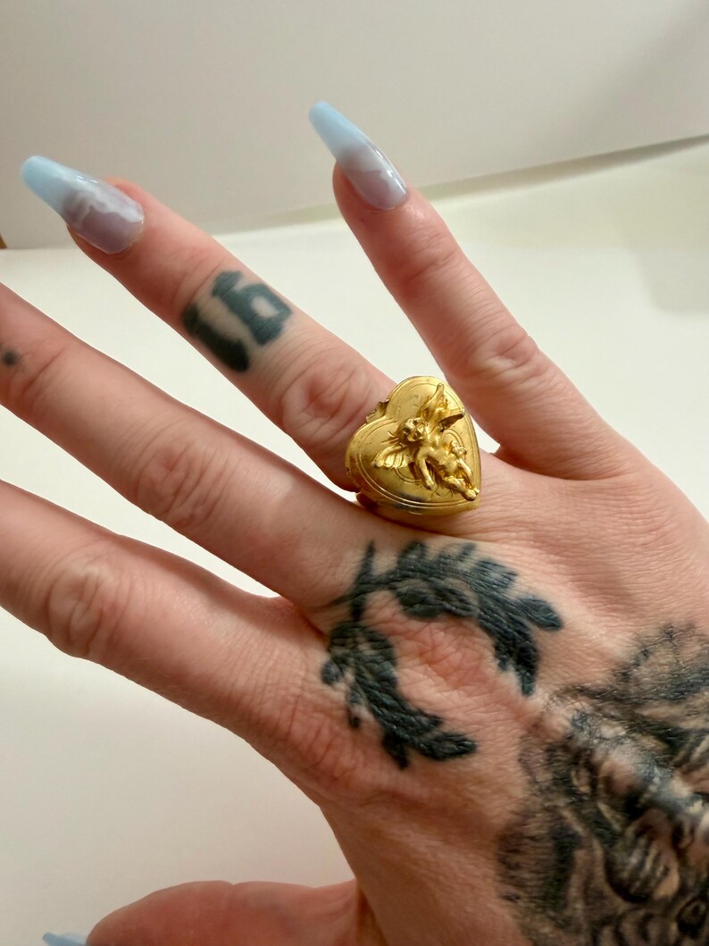 May include: A gold-colored heart-shaped ring with an embossed cherub design. The ring is worn on a finger. The ring's design features a heart shape with a cherub on top. The ring is a piece of jewelry.