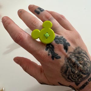 May include: A lime green button ring shaped like Mickey Mouse. The ring has a green cross-stitch detail in the centre. A playful and unique accessory.