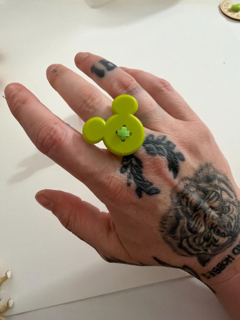 May include: A lime green button ring shaped like Mickey Mouse. The ring has a green cross-stitch detail in the centre. A playful and unique accessory.