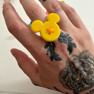 May include: A yellow button ring in the shape of a cartoon mouse. The ring features a large, round button with two smaller circular buttons for ears. The button is stitched with orange thread. Other colourful rings are in the background.