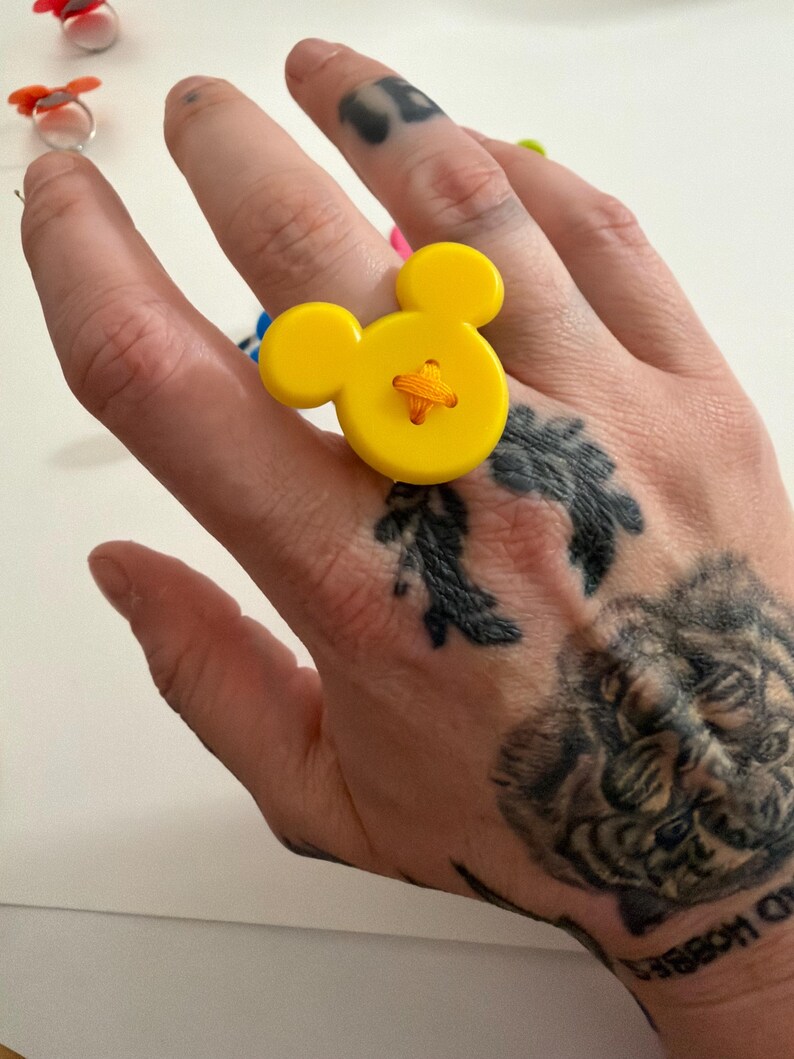 May include: A yellow button ring in the shape of a cartoon mouse. The ring features a large, round button with two smaller circular buttons for ears. The button is stitched with orange thread. Other colourful rings are in the background.
