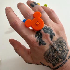 May include: An orange button ring shaped like Mickey Mouse. The ring is made from a large orange button with cross-stitch detail. A playful and fun accessory.