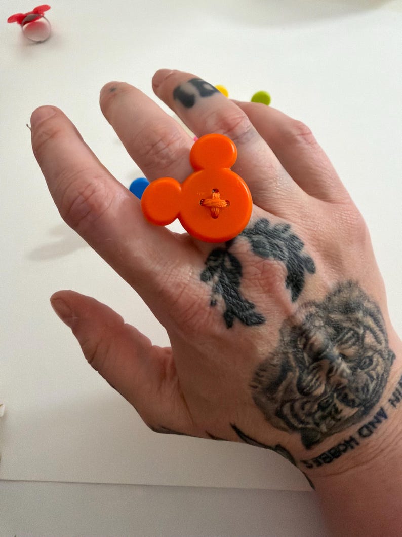 May include: An orange button ring shaped like Mickey Mouse. The ring is made from a large orange button with cross-stitch detail. A playful and fun accessory.