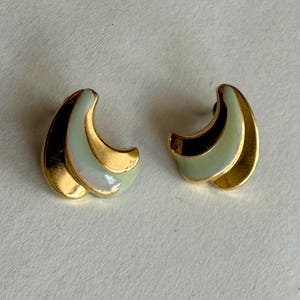 May include: A pair of gold-toned earrings with a curved design. Each earring features a gold outer edge, a central section of pale green, and a dark brown accent. The earrings are set against a white background.