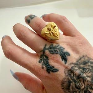 May include: A gold-colored ring in the shape of a heart, featuring a detailed relief of a cherub. The ring is worn on a finger. The ring's design is intricate, with fine details visible on the cherub and the heart's surface.