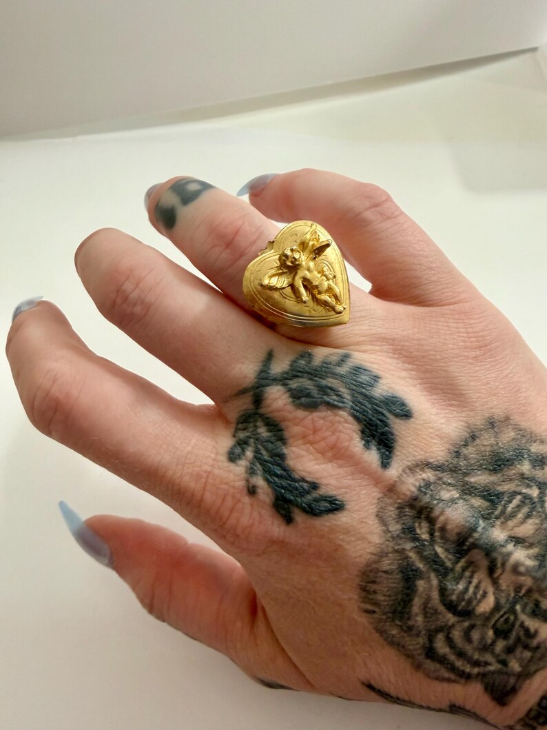 May include: A gold-colored ring in the shape of a heart, featuring a detailed relief of a cherub. The ring is worn on a finger. The ring's design is intricate, with fine details visible on the cherub and the heart's surface.