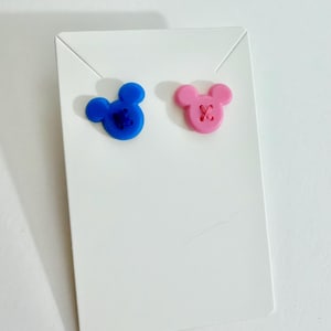 May include: A pair of stud earrings in the shape of a mouse head, one blue and one pink, each with a stitched 'X' detail. The earrings are displayed on a white earring card.