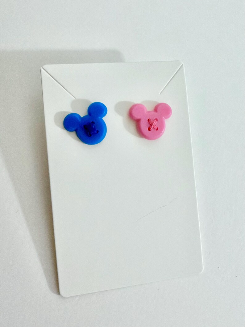 May include: A pair of stud earrings in the shape of a mouse head, one blue and one pink, each with a stitched 'X' detail. The earrings are displayed on a white earring card.