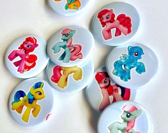 Può includere: Una collezione di otto spille rotonde, ognuna con un diverso personaggio pony colorato. Le spille hanno uno sfondo azzurro e mostrano vari pony nelle tonalità del rosa, giallo, blu e verde con criniere e code distintive.