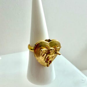 May include: A gold-colored ring featuring a heart-shaped design with an embossed cherub figure. The ring is displayed on a white, cone-shaped stand, showcasing its intricate details and metallic finish. The ring's band is a simple, smooth design.