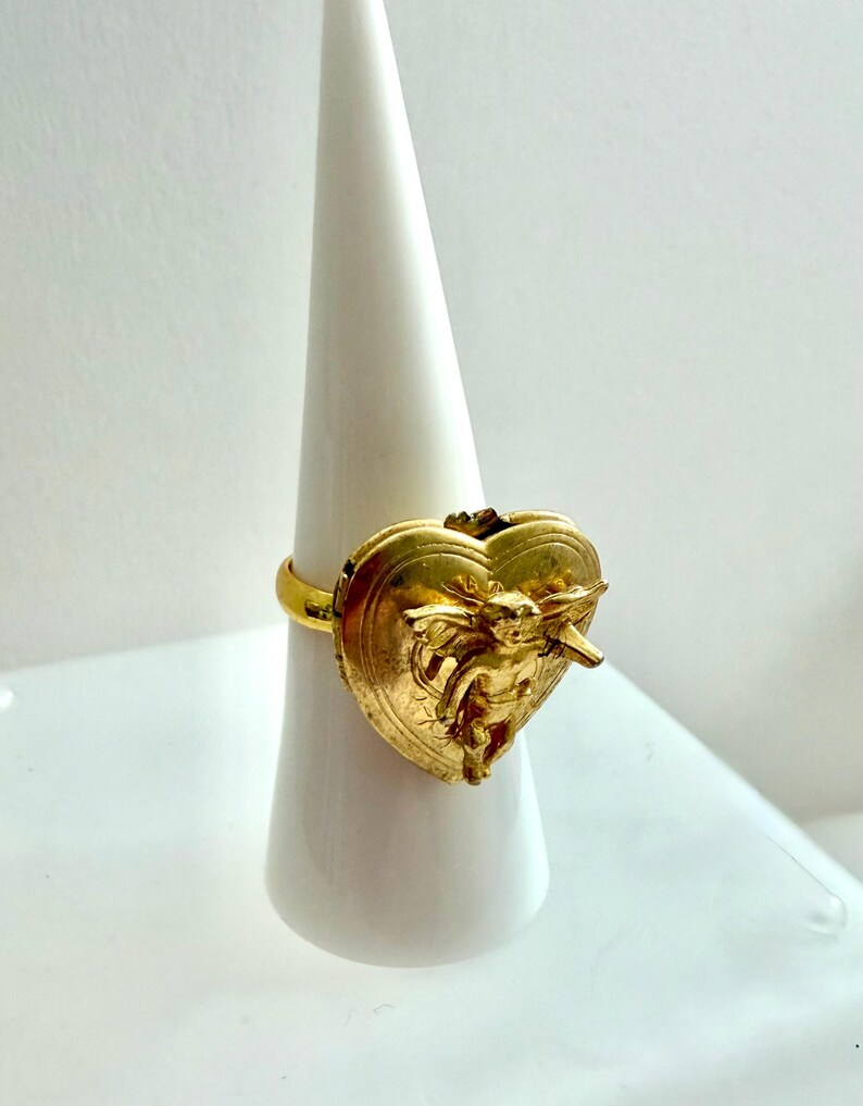 May include: A gold-colored ring featuring a heart-shaped design with an embossed cherub figure. The ring is displayed on a white, cone-shaped stand, showcasing its intricate details and metallic finish. The ring's band is a simple, smooth design.