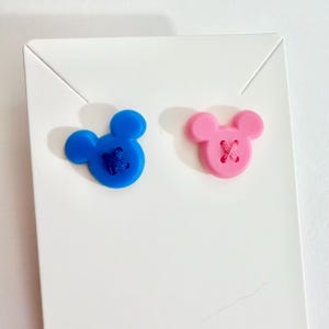 May include: Two Mickey Mouse-shaped buttons. One is blue with blue stitching, and the other is pink with pink stitching. The buttons are on a white card.