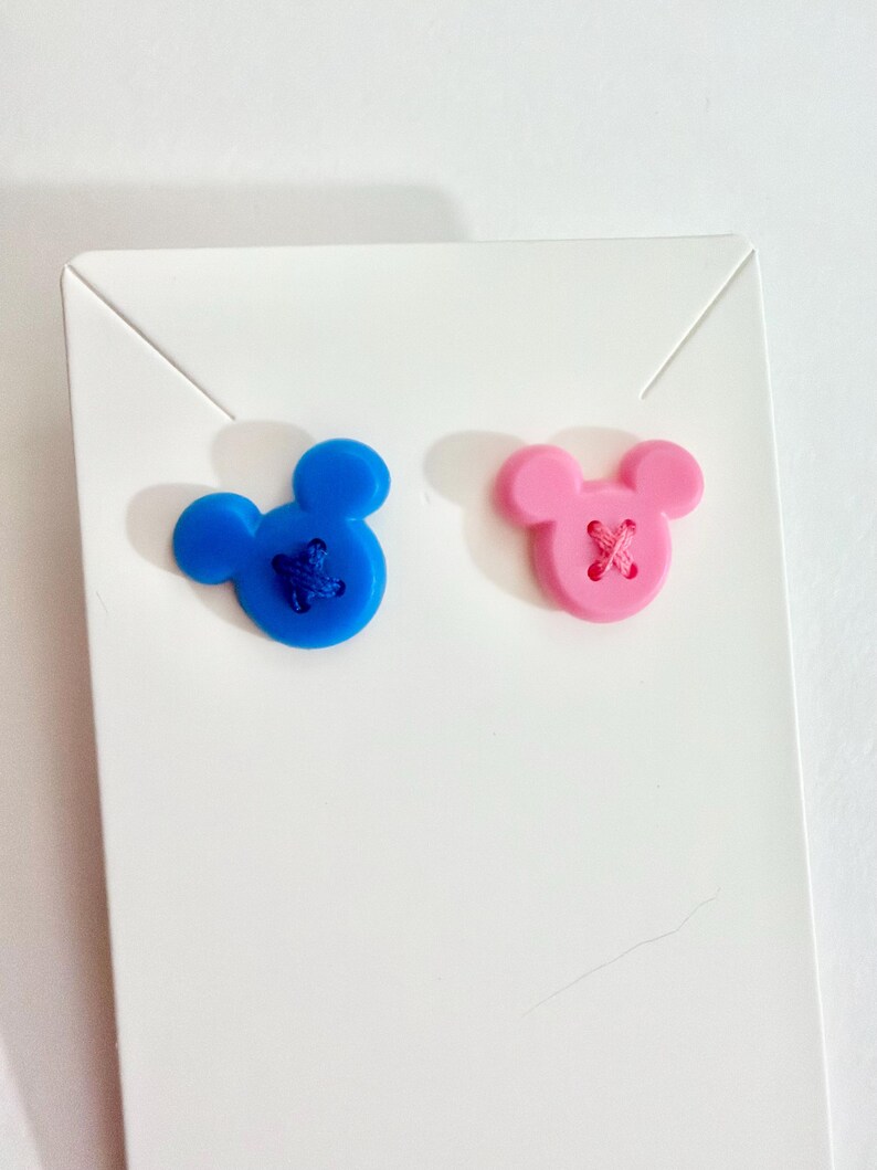 May include: Two Mickey Mouse-shaped buttons. One is blue with blue stitching, and the other is pink with pink stitching. The buttons are on a white card.