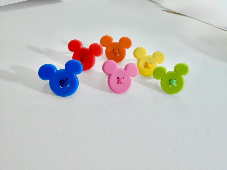 May include: Six colourful buttons in the shape of a mouse's head. The buttons are in blue, red, orange, pink, yellow, and green. Each button has contrasting thread sewn through the centre.