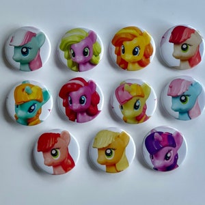 May include: A collection of twelve round buttons featuring colorful cartoon pony characters. Each button displays a different pony with varying colors, including pink, yellow, blue, and purple, set against a white background. The buttons are arranged in a circular pattern.