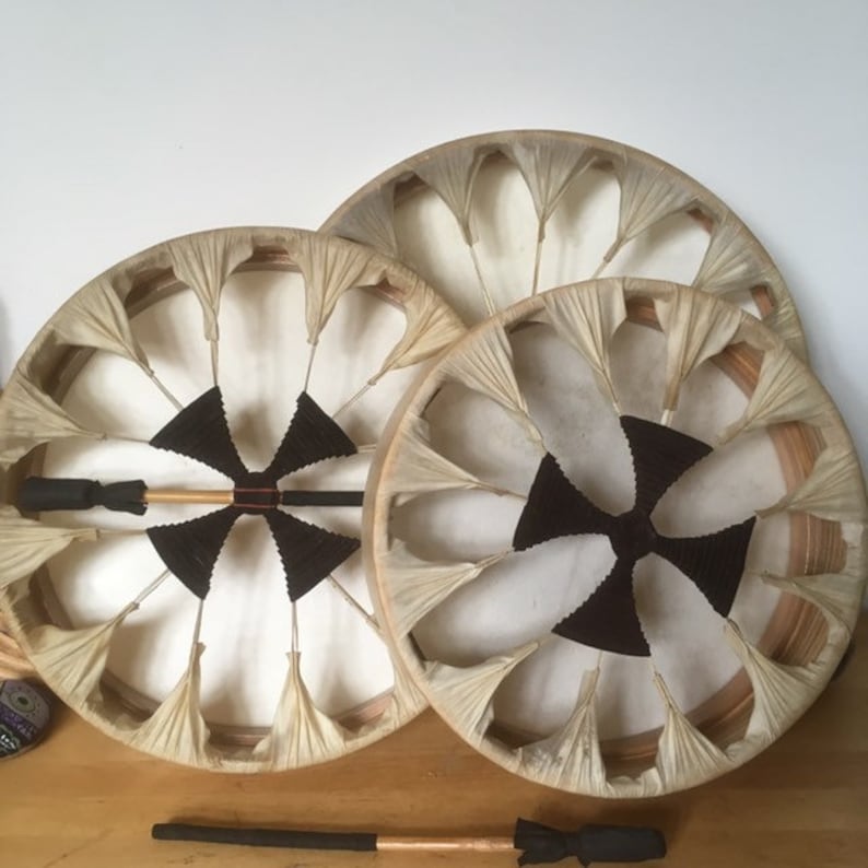 May include: Three round, handmade drums with natural-toned skins and wooden frames. Each drum features a central black design and a unique fan-like pattern around the edges. A drumstick with a black handle and copper end rests below.