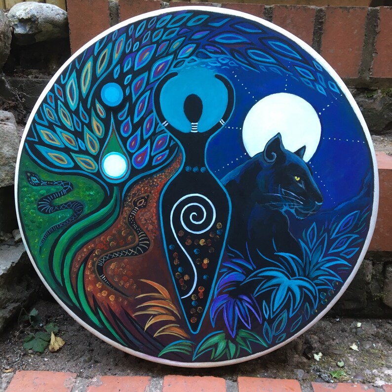 May include: A circular, hand-painted artwork depicting a central female figure with raised arms. The artwork is split into three sections: a tree with snakes, the figure, and a panther under a full moon. The colour palette includes blues, greens, browns, and white.