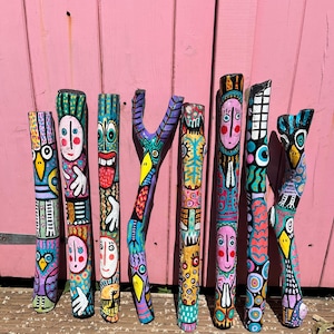 May include: Eight wooden sticks painted with vibrant, multicolored patterns and stylized faces. Each stick features unique designs including birds, human-like figures, and abstract shapes in bright shades of blue, pink, yellow, and black.
