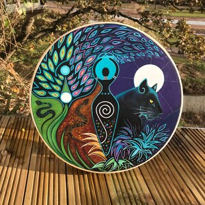 May include: A circular, hand-painted artwork depicting a black panther, a full moon, and stylized figures. The design uses bright colours, including blues, greens, and browns, with a wooden frame. The artwork has a spiritual or mystical theme.