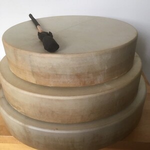 May include: Three stacked, round frame drums with natural-toned drumheads. A drumstick with a black head rests on the top drum. The drums are of varying sizes, creating a tiered effect. The drums are likely used for music or sound healing.