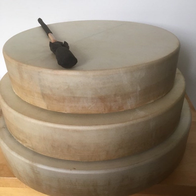 May include: Three stacked, round frame drums with natural-toned drumheads. A drumstick with a black head rests on the top drum. The drums are of varying sizes, creating a tiered effect. The drums are likely used for music or sound healing.
