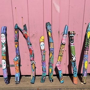 Könnte beinhalten: Eine Sammlung von acht handbemalten Holzstücken, jedes mit einem einzigartigen Design. Die Stücke sind mit leuchtenden Farben bemalt und zeigen Gesichter, Vögel und geometrische Muster. Sie stehen aufrecht vor einem rosa Holzgrund.