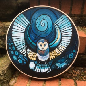 May include: A round, hand-painted artwork featuring a stylized owl with outstretched wings. The owl is depicted in shades of blue, brown, and white against a dark blue background with a spiral design. A small blue mouse is also visible.