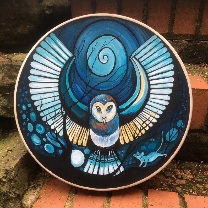 May include: A round, hand-painted artwork featuring a stylized owl with outstretched wings. The owl is depicted in shades of blue, brown, and white against a dark blue background with a spiral design. A small blue mouse is also visible.