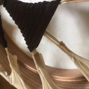 May include: Close-up of a decorative object featuring dark brown, triangular fabric elements attached to thin, beige stems. The stems are connected to a copper-colored circular frame, creating a layered, textured design. The background is a soft, off-white color.