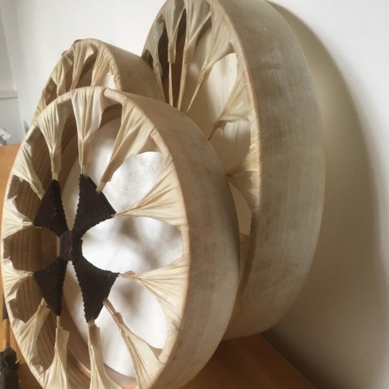 May include: Three circular drums with wooden frames and off-white drumheads. The frames have a unique design with radiating fabric accents. The centre of each drum has a dark brown, star-like pattern. The drums are handmade.