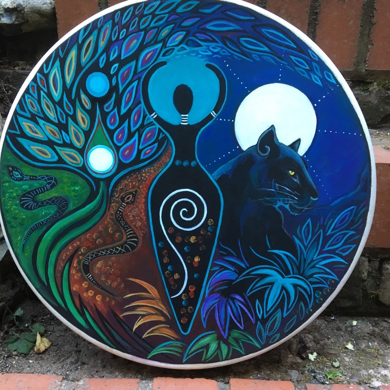 May include: Circular painting depicting a central female figure, a black panther, and a full moon. The artwork features a tree, snakes, and various plant life in shades of blue, green, brown, and black. The painting has a white border.