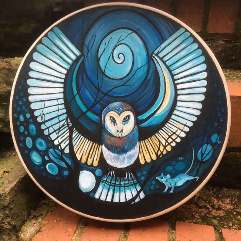 May include: A round, hand-painted wooden art piece featuring a stylized owl with outstretched wings. The owl is depicted in shades of blue, brown, and yellow against a dark blue background with a spiral design. A small blue mouse is also visible.