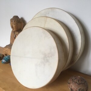 May include: Three circular drums in shades of cream are stacked, with a wooden Buddha statue and decorative stones in the background. The drums have a natural, textured appearance, suggesting a handmade quality. The image evokes a sense of spirituality and music.