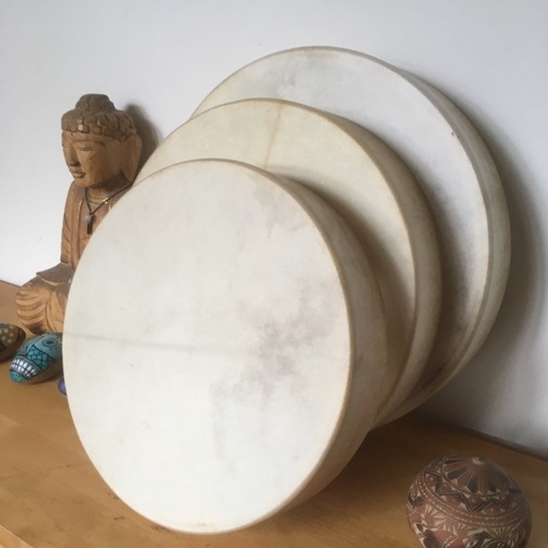 May include: Three circular drums in shades of cream are stacked, with a wooden Buddha statue and decorative stones in the background. The drums have a natural, textured appearance, suggesting a handmade quality. The image evokes a sense of spirituality and music.