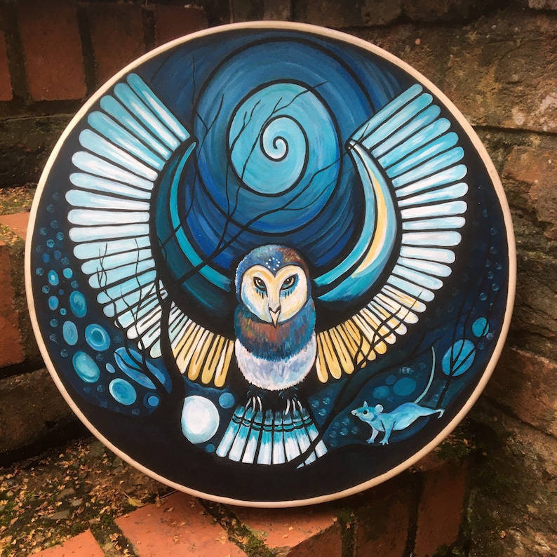 May include: A round, hand-painted artwork featuring a stylized owl with outstretched wings. The owl is depicted in shades of blue, brown, and yellow against a dark blue background with a swirling design. A small blue mouse is also present. The artwork is framed with a light-colored wooden edge.