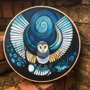 May include: Round wall art featuring a stylized owl with outstretched wings, painted in shades of blue, white, and yellow against a dark background. A small blue mouse is depicted at the bottom. The artwork is framed with a light-colored wooden border.