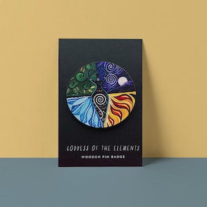 Op de afbeelding: Een houten pin met een cirkelvormig ontwerp met een godin verdeeld in vier secties die de elementen vertegenwoordigen. De secties omvatten groen gebladerte, blauw water, geel vuur en een nachthemel met een maan. Tekst: "Goddess of the Elements".