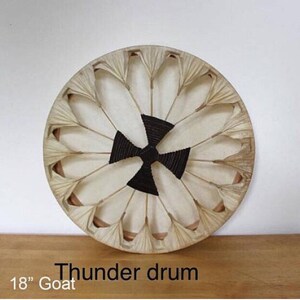 May include: A round thunder drum made from goat skin, measuring 45.7 cm in diameter. The drum has a central black woven pattern and is surrounded by white petal-shaped elements. The frame is a natural beige color.