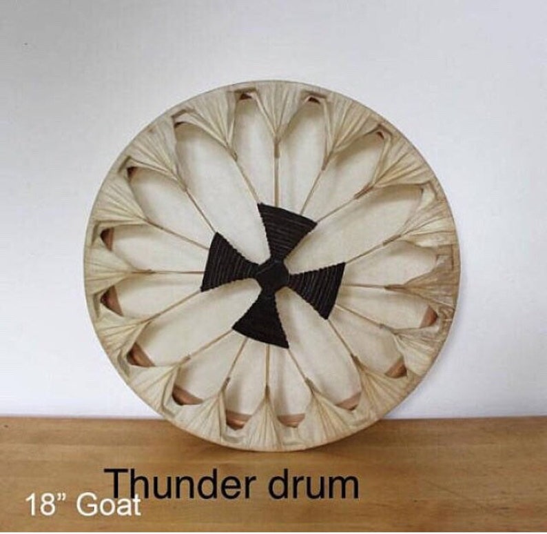 May include: A round thunder drum made from goat skin, measuring 45.7 cm in diameter. The drum has a central black woven pattern and is surrounded by white petal-shaped elements. The frame is a natural beige color.