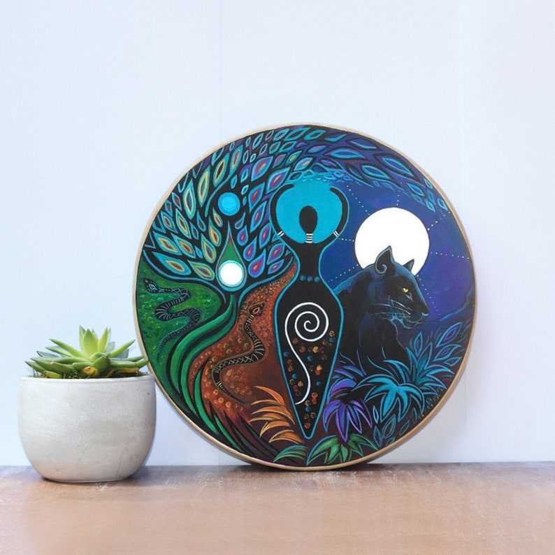 May include: A circular painting with a central female figure, a black panther, and a tree with snakes, set against a night sky with a full moon and foliage. The artwork is displayed on a wooden surface. The painting uses a vibrant colour palette.